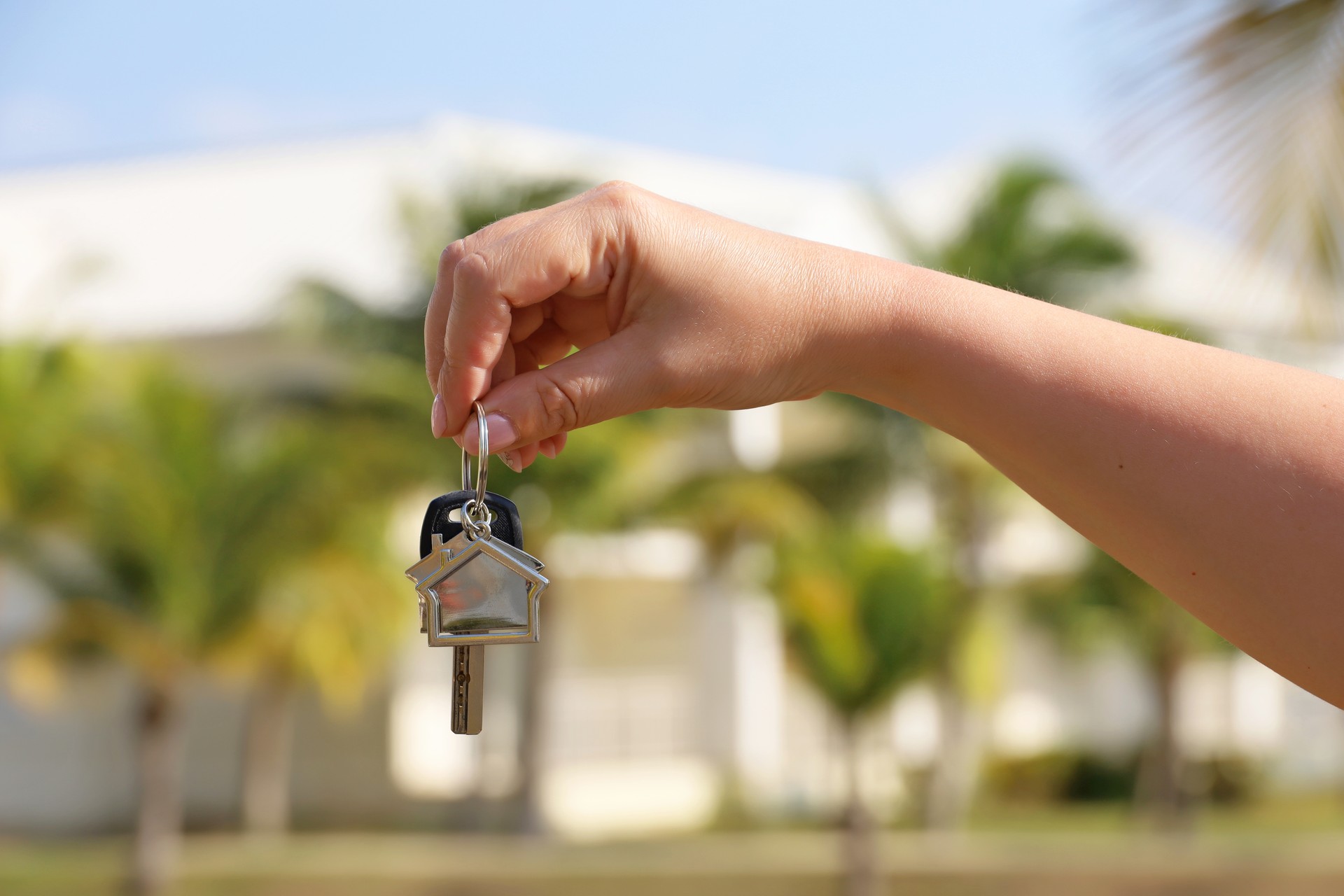Real estate agent, home keys in female hand on background of house surrounded by palm trees
